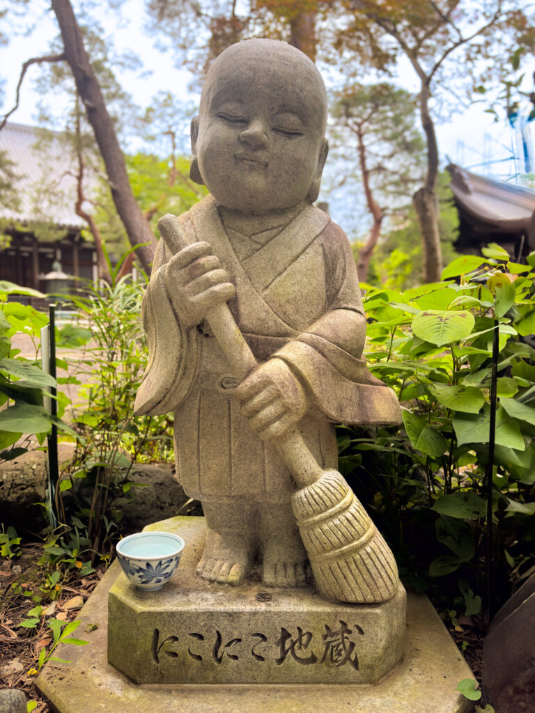 Smiling Jizo statue with broom surrounded by greenery
